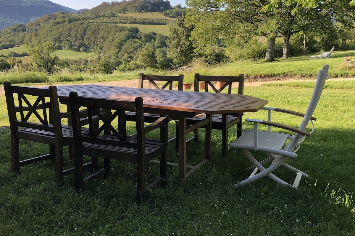 A wooden table and chairs set on a grassy field with a view of rolling hills and trees.