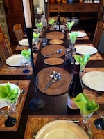 Una larga mesa de comedor dispuesta con platos, cubiertos y copas de vino, con individuales tejidos y sujetavasos verdes.
