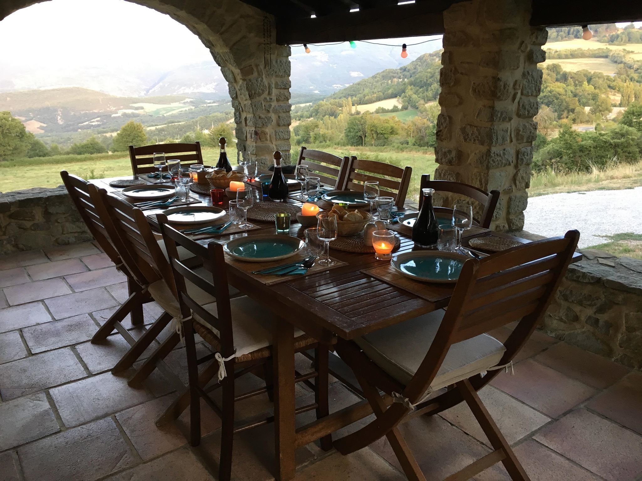 Un área de comedor al aire libre con pilares de piedra, mesas y sillas de madera, frente a un hermoso paisaje de montañas.