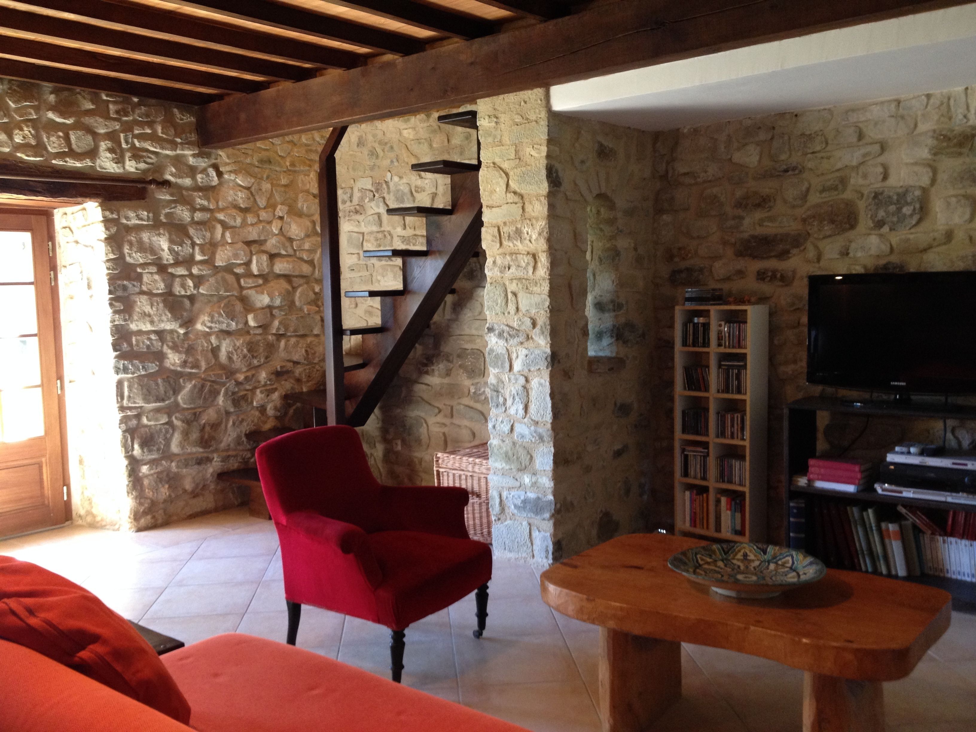Una habitación con paredes de piedra, una escalera de madera, un sillón rojo, una mesa de madera y una estantería con libros y un televisor.