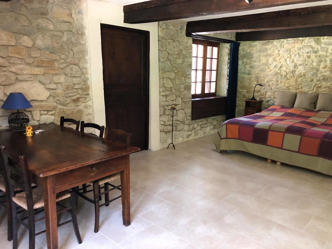 Una habitación con pared de piedra, puerta de madera, una mesa de comedor con sillas, cama con colcha colorida y ventana con vistas.