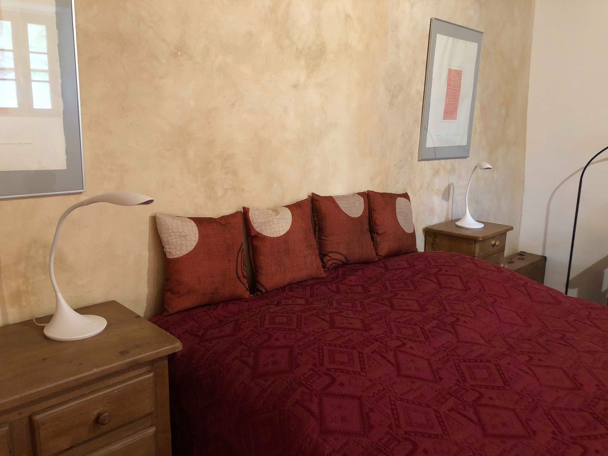 A bedroom with a patterned red bedspread, a wooden nightstand with a white lamp, and a framed picture on the wall.