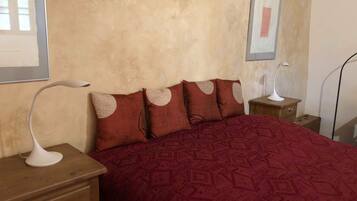 A bedroom with a patterned red bedspread, a wooden nightstand with a white lamp, and a framed picture on the wall.