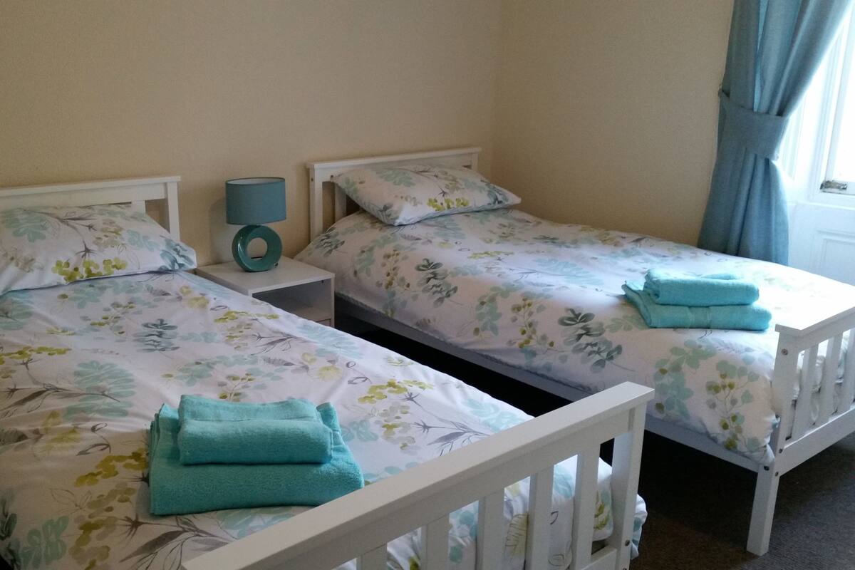 Two single beds with floral bedding, a bedside table with a lamp, and a window with curtains.