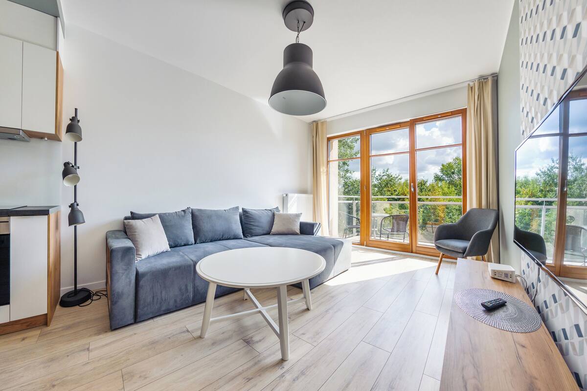 A modern living room with a grey sofa, a round white coffee table, and a large window with a view of greenery.