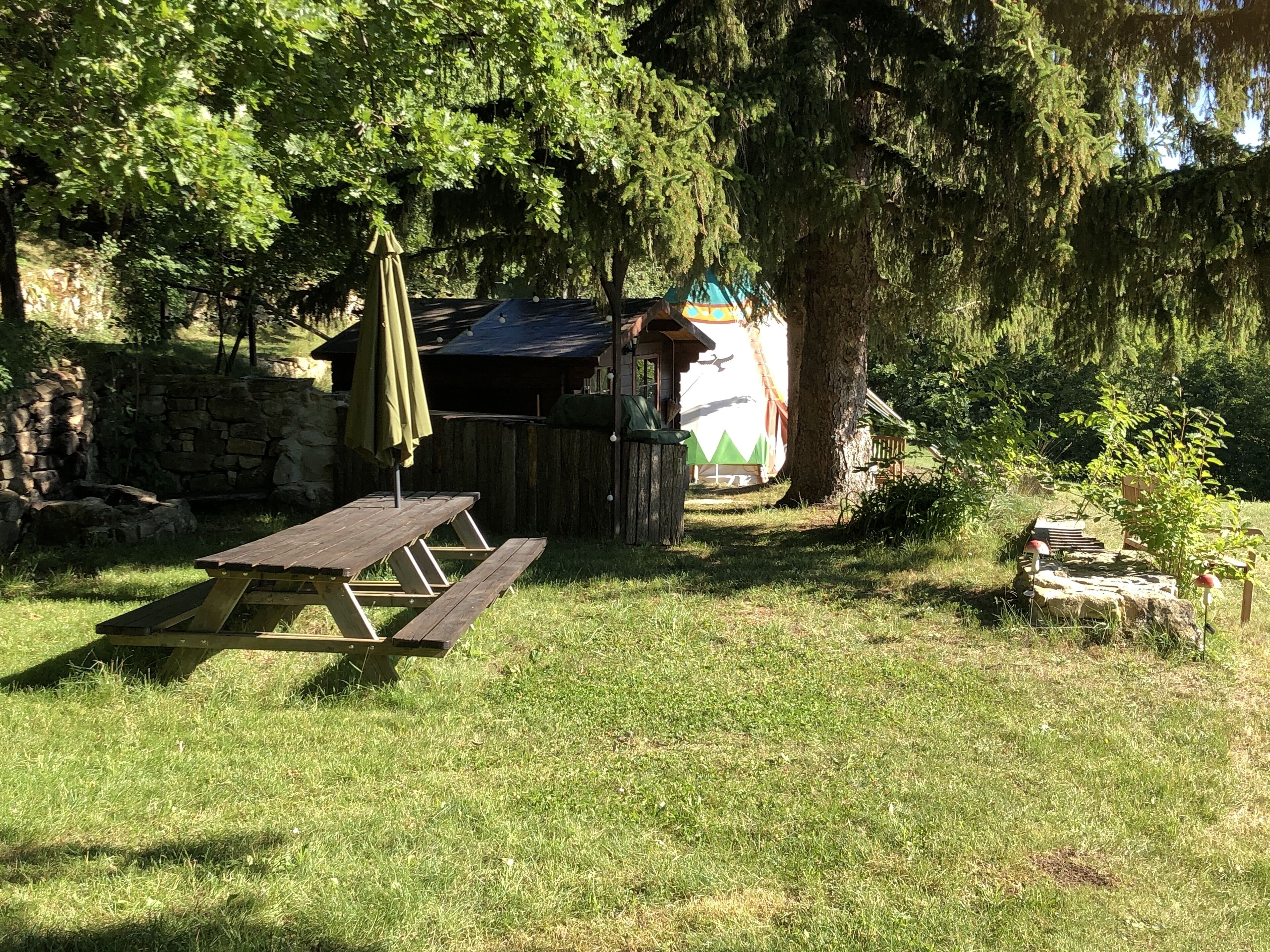 A picnic table with a green umbrella, a rustic wooden structure, and a stone wall in a grassy area with trees.