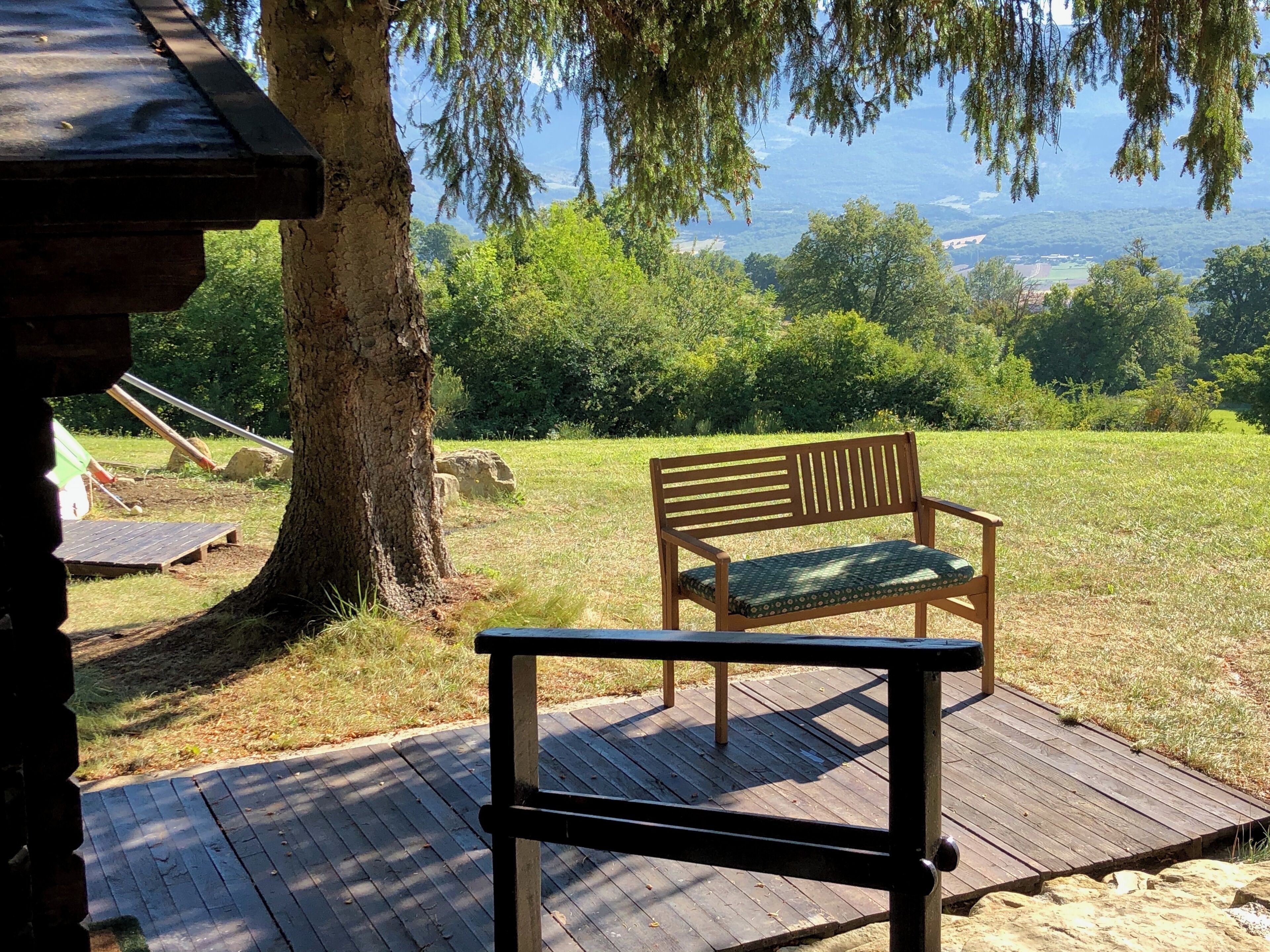 A wooden bench with a cushion on a wooden deck overlooking a grassy area with trees and a distant view of mountains.