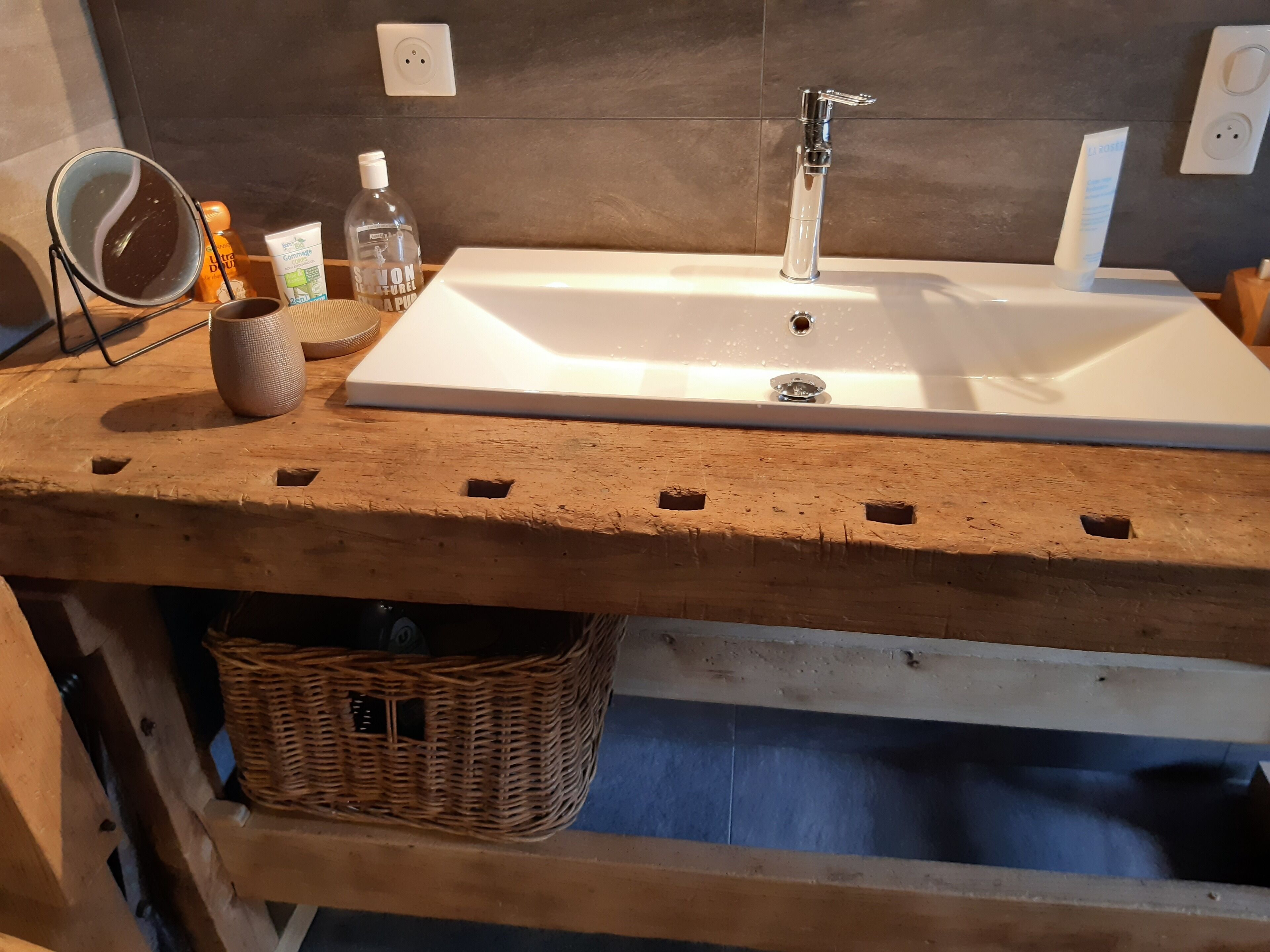 A bathroom with a wooden vanity, a white sink, a mirror, a bottle of shampoo, and a soap dispenser.