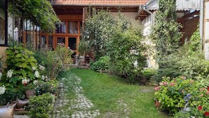 A cobblestone pathway leading to a house with a red-tiled roof, surrounded by lush greenery and flowering plants. - Un Soir d'Eté (Ernolsheim-Bruche)