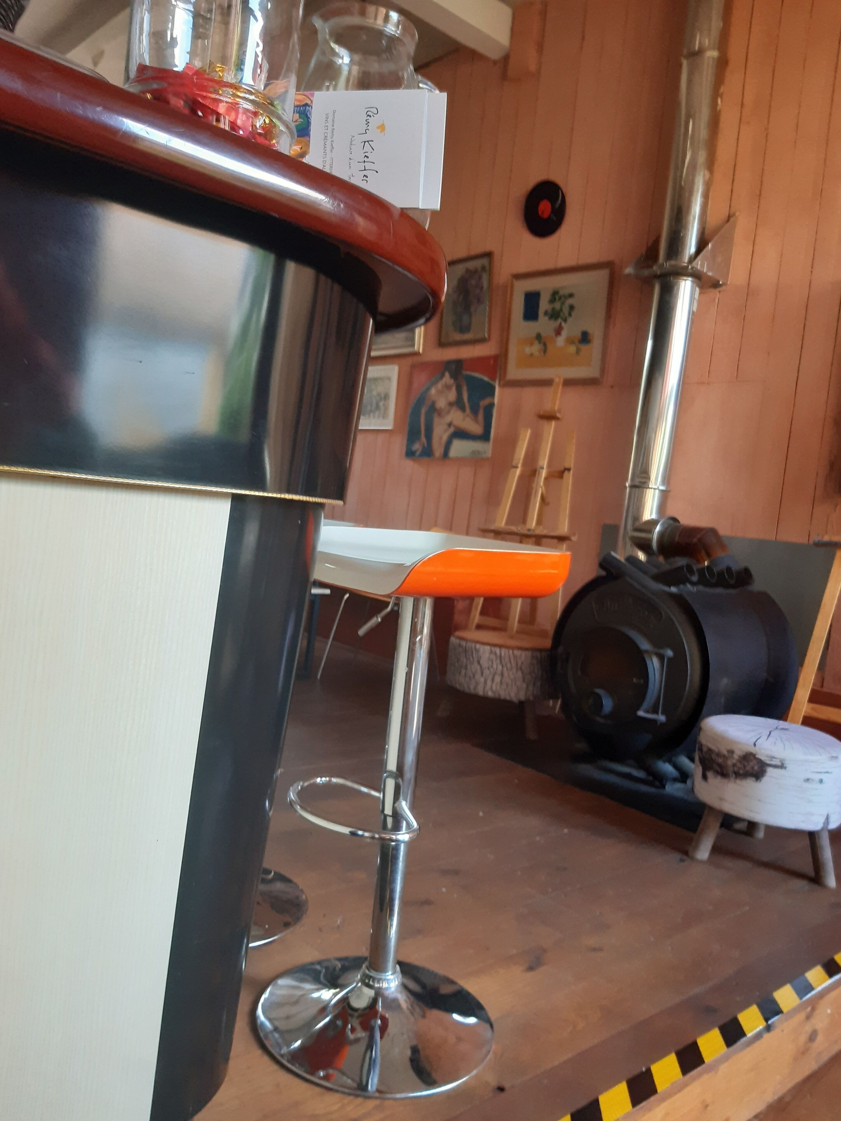 Un taburete con base cromada y asiento naranja, una barra de madera y una cafetera de estilo vintage al fondo.