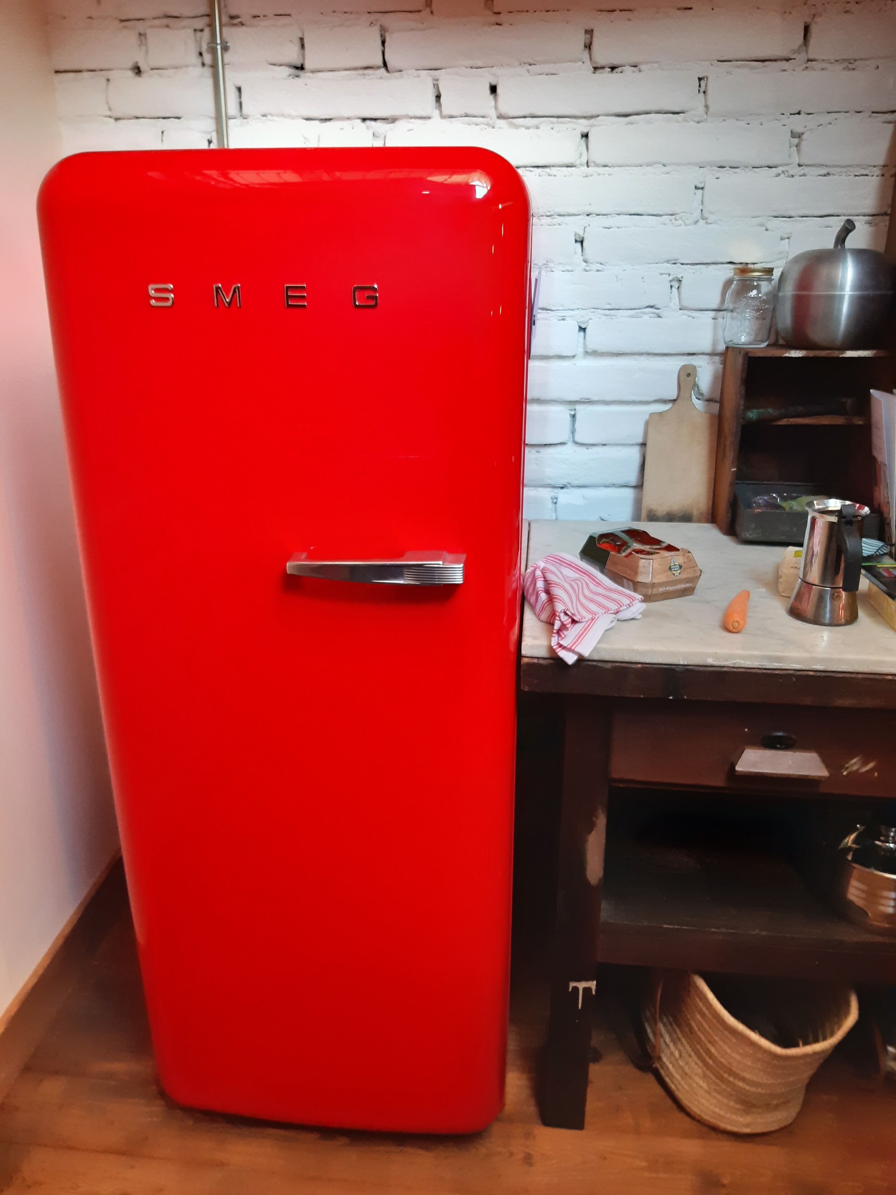 A red Smeg refrigerator in a kitchen with a white brick wall, wooden table, and various kitchen items.