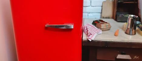 A red Smeg refrigerator in a kitchen with a white brick wall, wooden table, and various kitchen items.