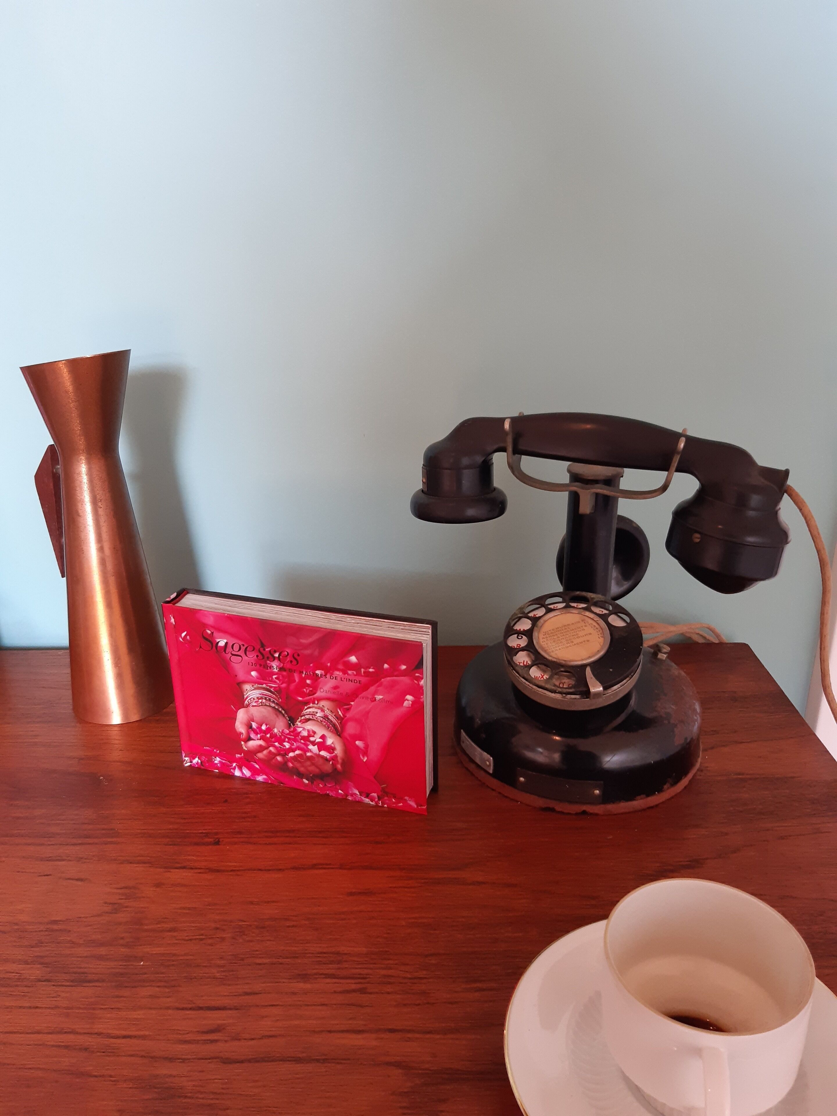 Een oude telefoon met draaiknoppen, een rood boek en een witte beker op een houten tafel.