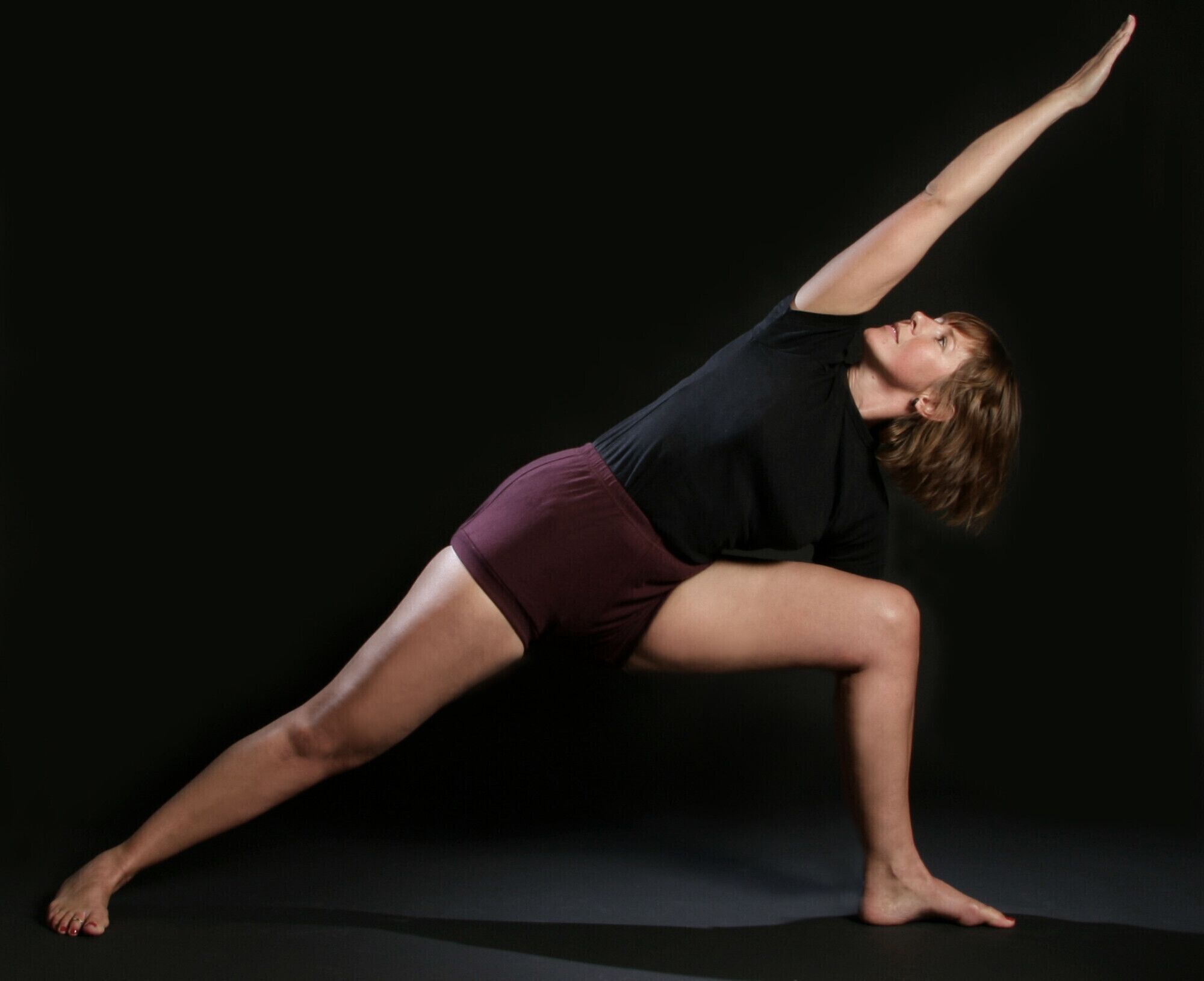 A person in a yoga pose on a dark floor with a plain background.
