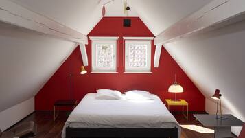 A bedroom with a sloped ceiling, two windows with grid patterns, a bed with white bedding, a bedside table with a lamp, and a small table with a lamp.