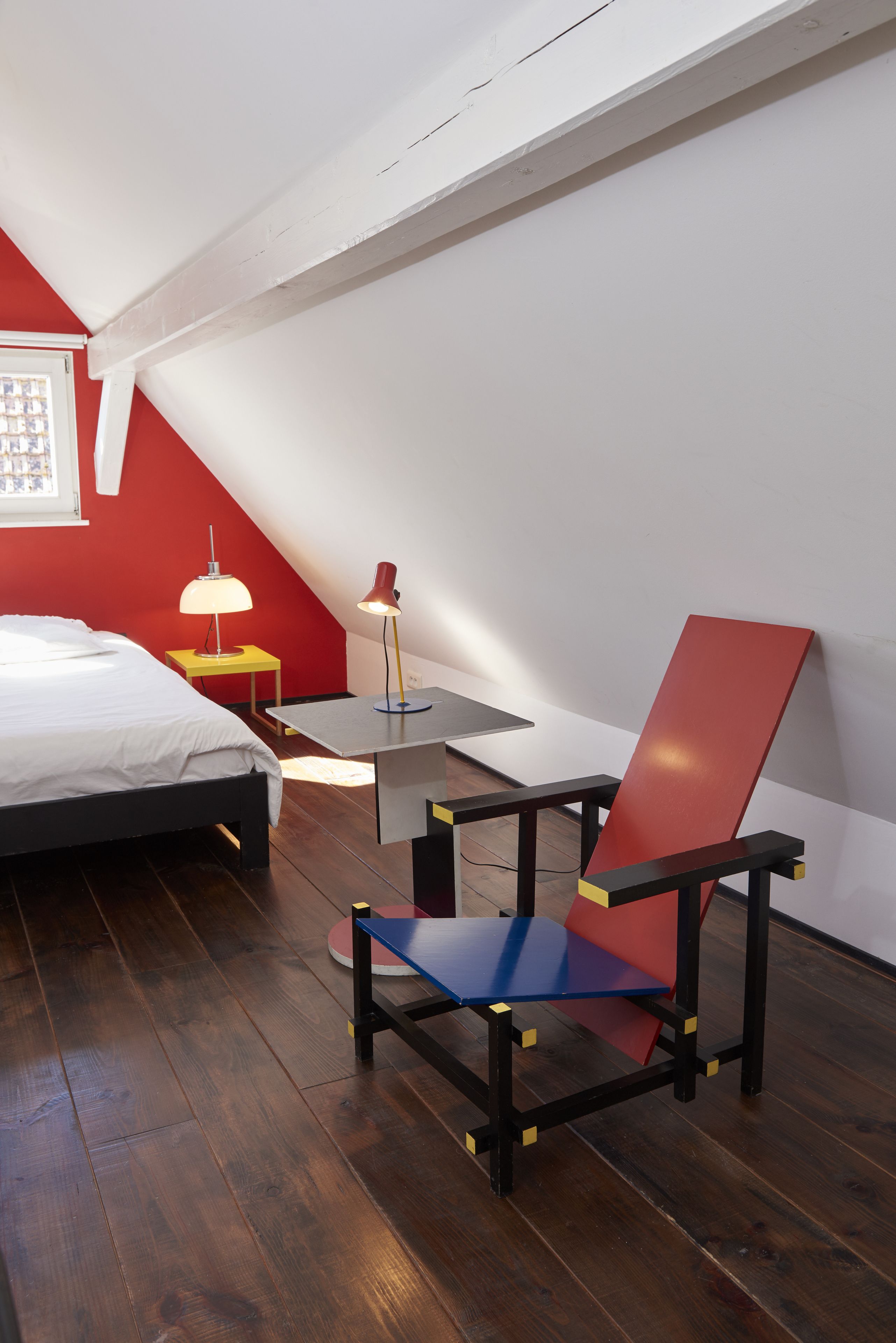 Una habitación con una cama, una silla roja y azul, una mesita y dos lámparas.