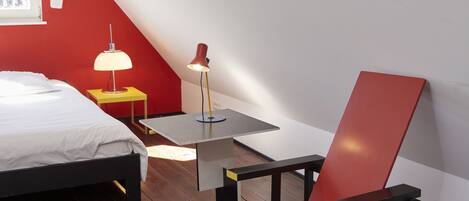 Una habitación con una cama, una silla roja y azul, una mesita y dos lámparas.