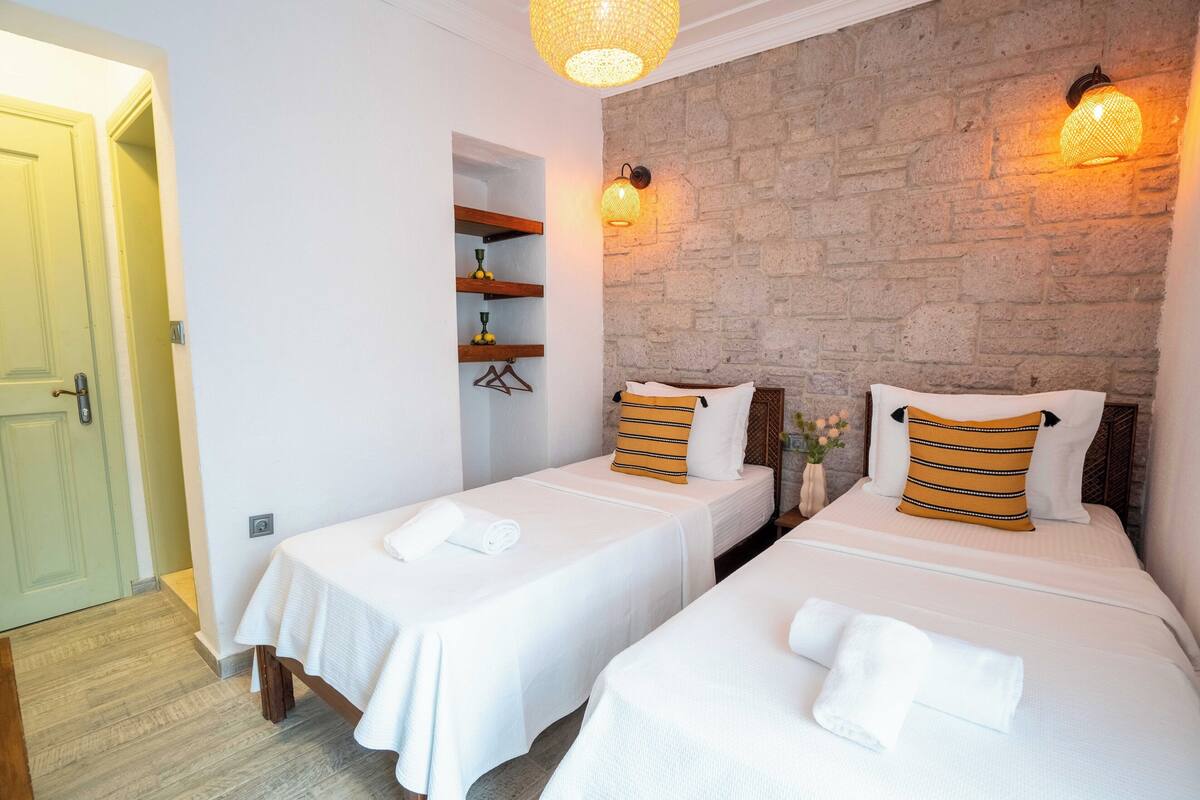 A hotel room with two single beds, a wooden door, a stone wall, and a shelf with items.