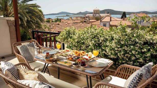Una terraza con muebles de mimbre, una mesa de comedor lista con comida y bebidas, y vistas al mar y edificios al fondo.