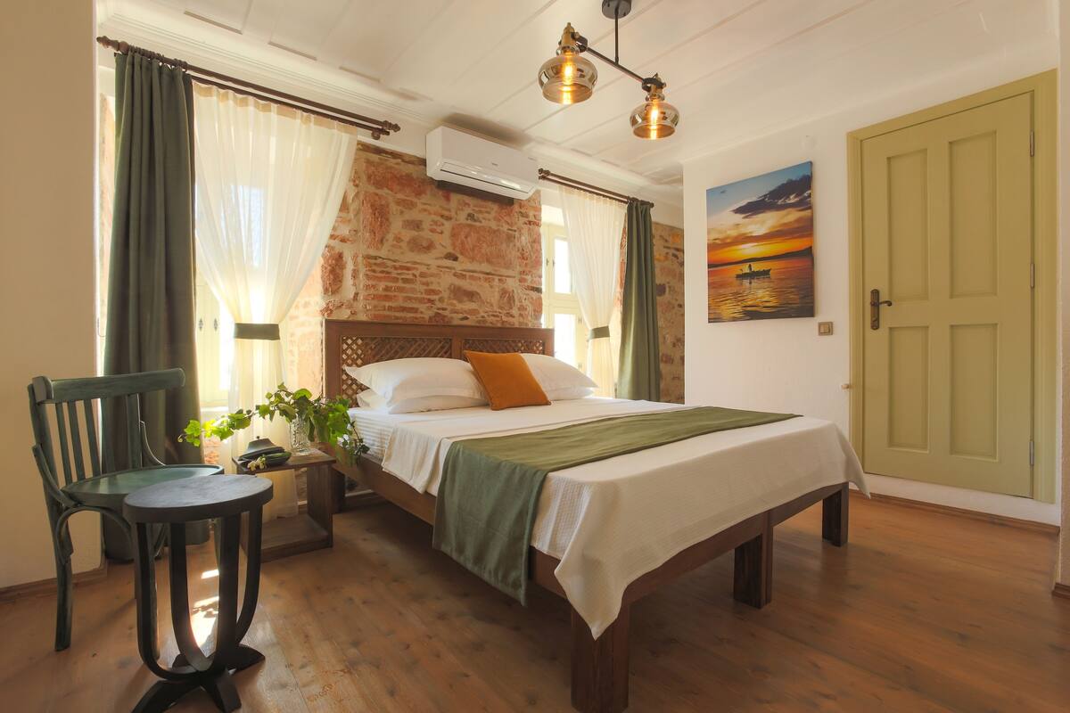 A bedroom with a wooden bed, a green chair, a small table, a painting of a sunset, and a green door.
