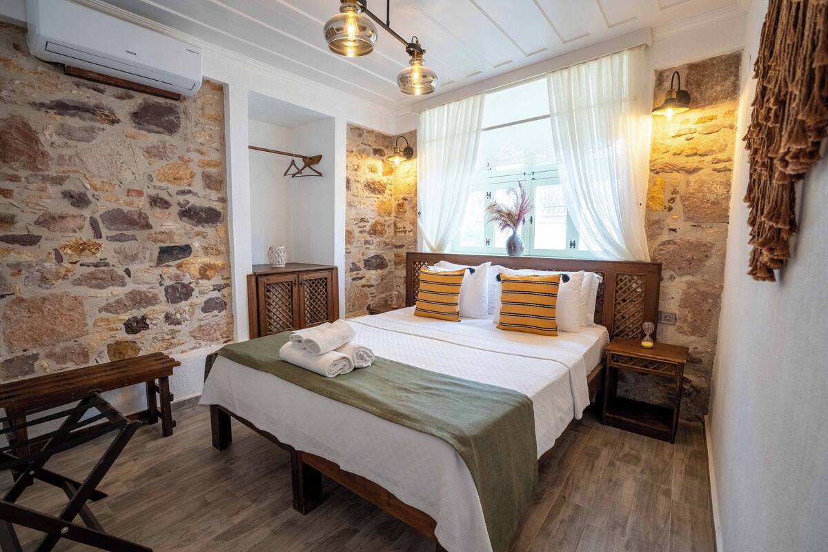 A cozy bedroom with a stone wall, a wooden bed, a bench, and a nightstand with a lamp.