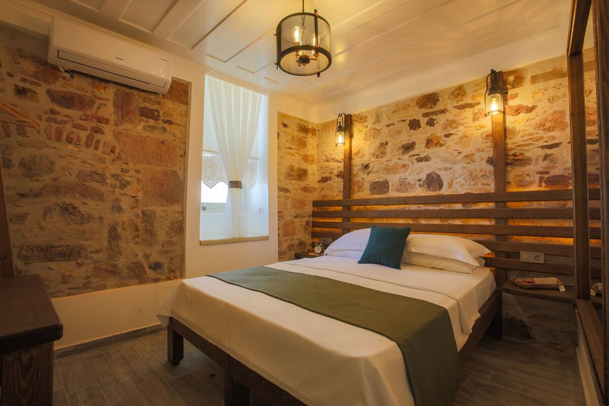 A bedroom with a stone wall, a wooden headboard, a bed with white linens and a green throw, a nightstand, and a window with sheer curtains.