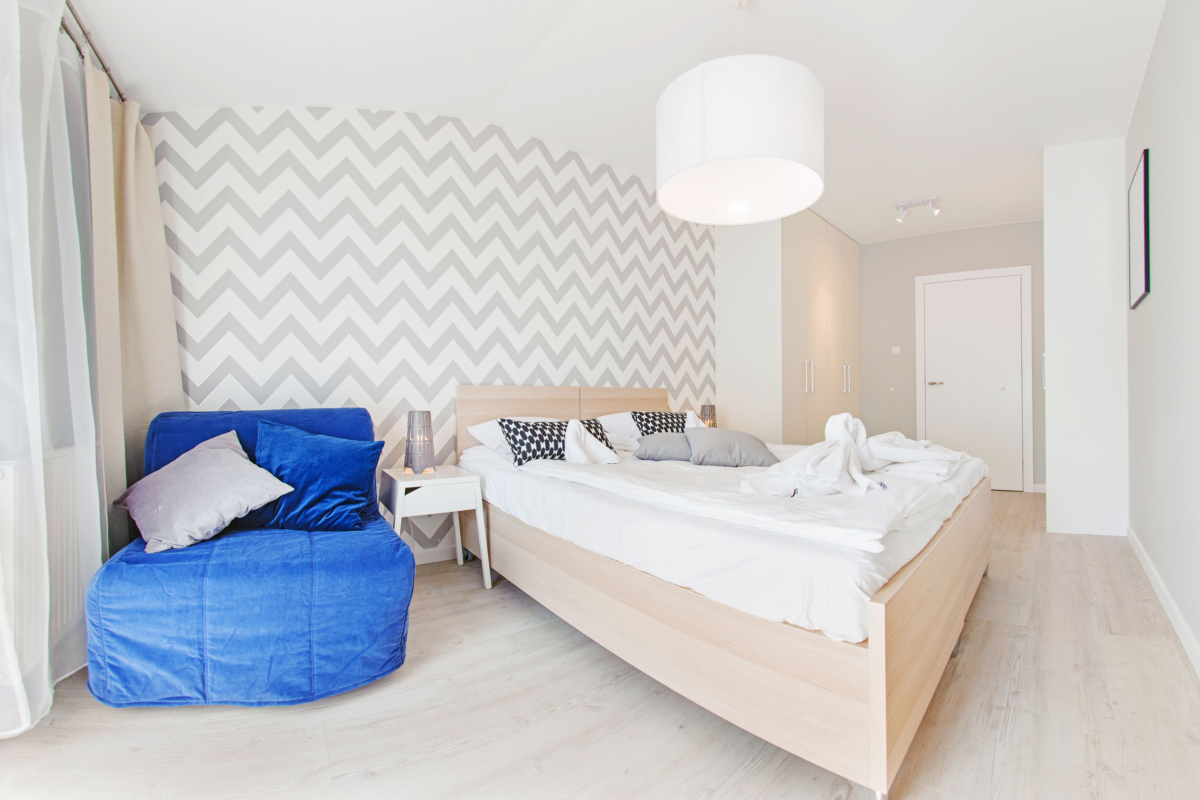 Une chambre moderne avec un lit, un fauteuil en forme de poire bleu, une table de chevet blanche et un mur orné d’un motif en zigzag.
