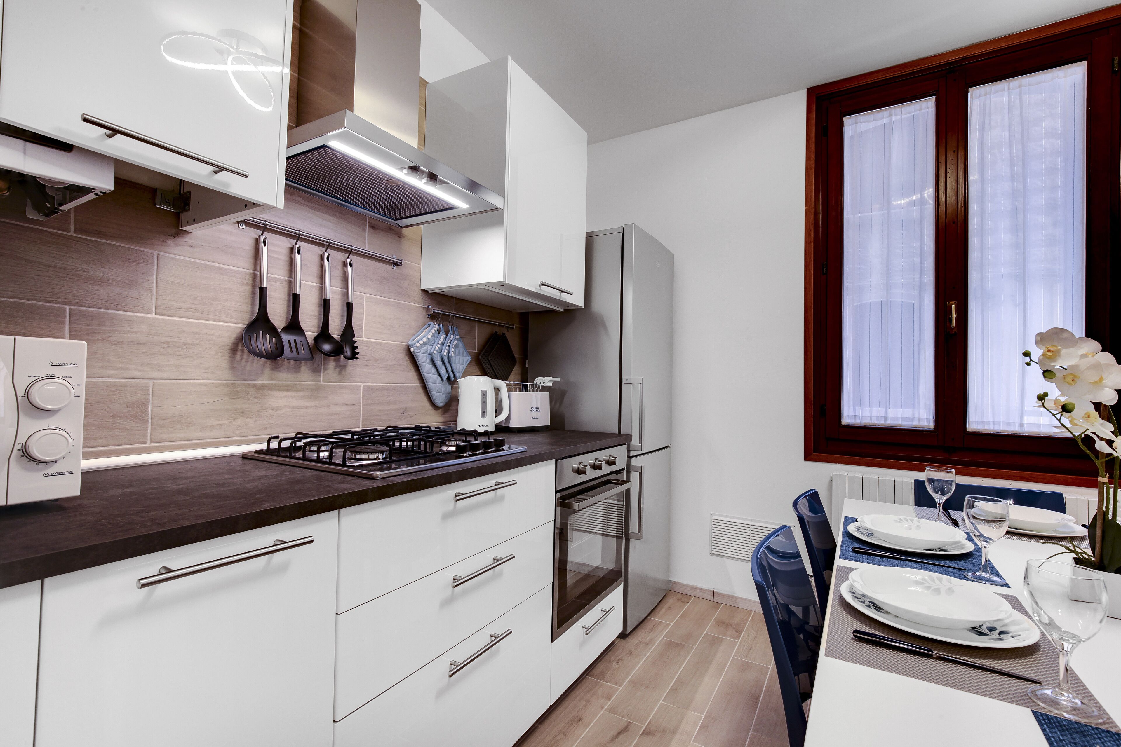 A modern kitchen with white cabinets, a gas stove, and a built-in oven. There is a dining area with a table set for two, and a window with curtains.