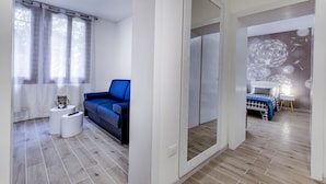Un salon moderne avec un canapé bleu, une table basse blanche et une petite table d’appoint. Un grand miroir orne le mur, et dans la pièce adjacente, on trouve un lit recouvert d’une couvre-lit à pois.
