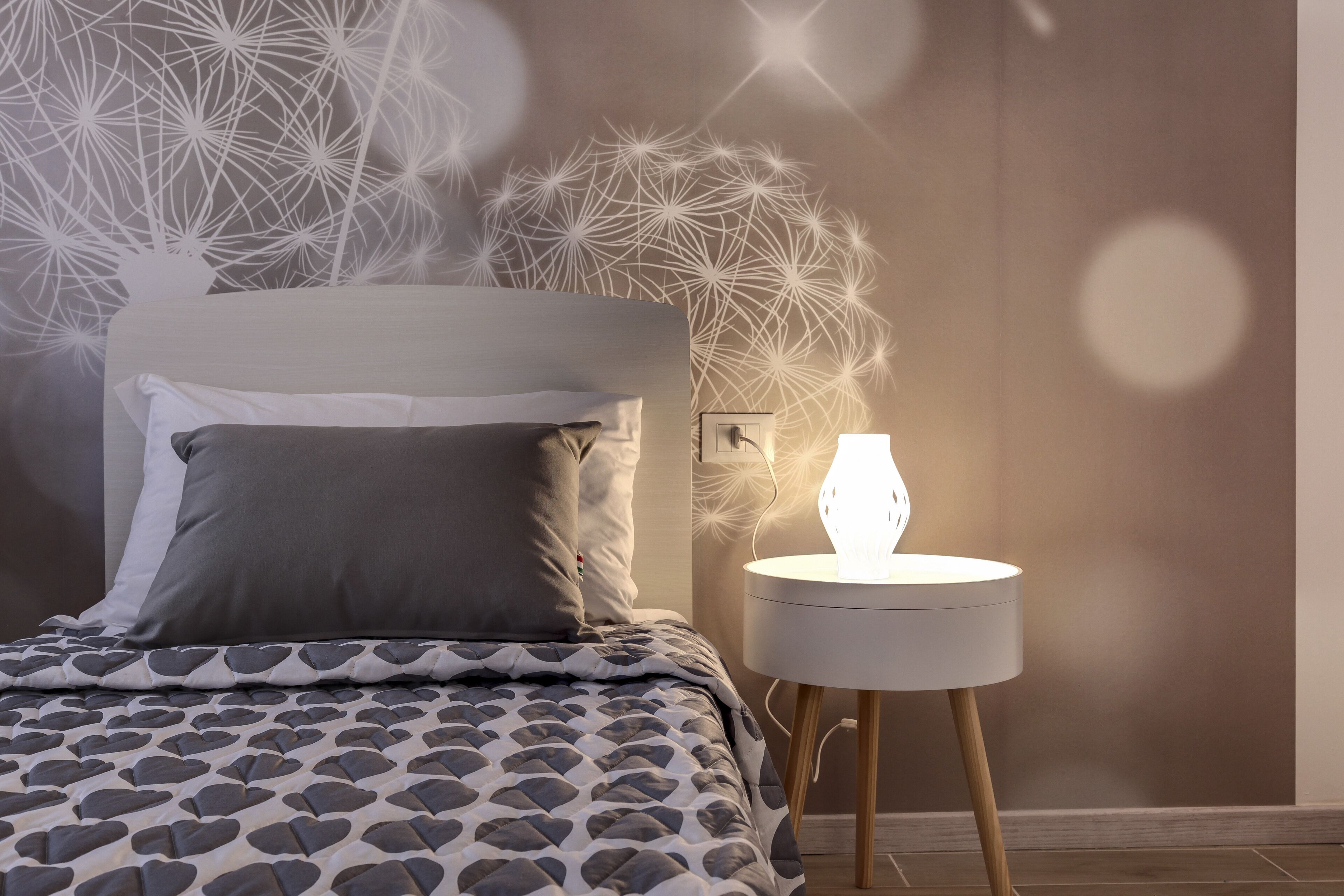 Ein ordentlich bezogenes Bett mit gemusterten Bettwäsche, ein Nachttisch mit Lampe und eine Wand mit geometrischem Muster.