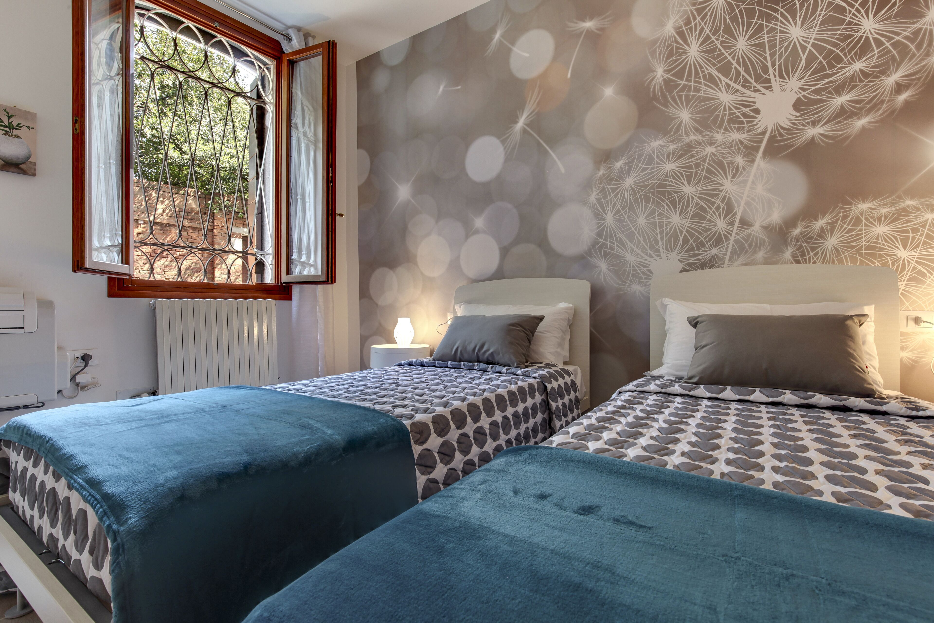Camera da letto con due letti, una finestra con tende, un termosifone e un muro decorato con un motivo a tarassaco.