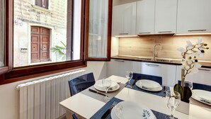 Un coin repas avec une table dressée pour deux personnes, une vue sur un bâtiment en pierre et une cuisine moderne aux meubles blancs.