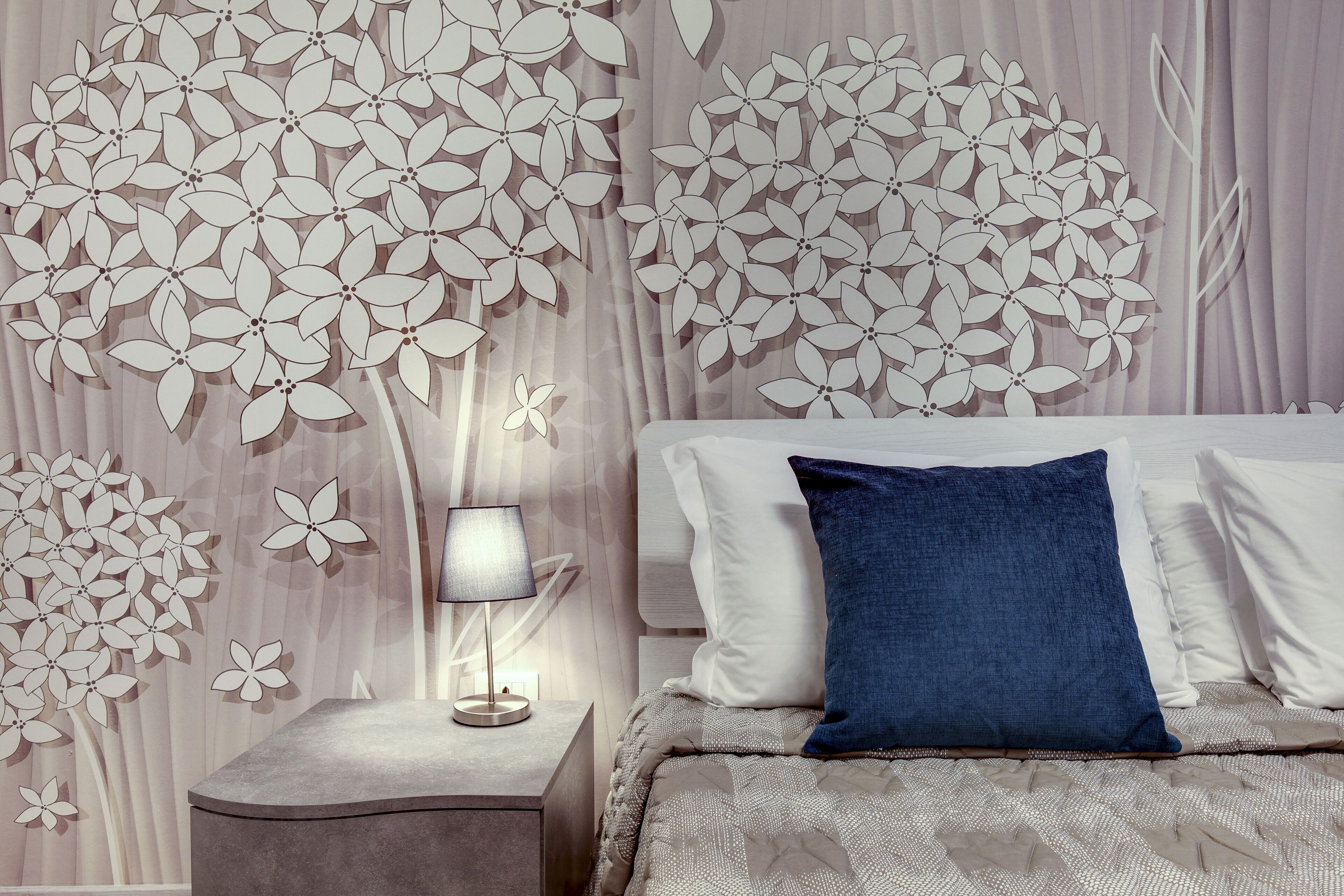 Ein Schlafzimmer mit Bett, Kissen, Nachttisch, Lampe und einer Wand mit floralem Muster.