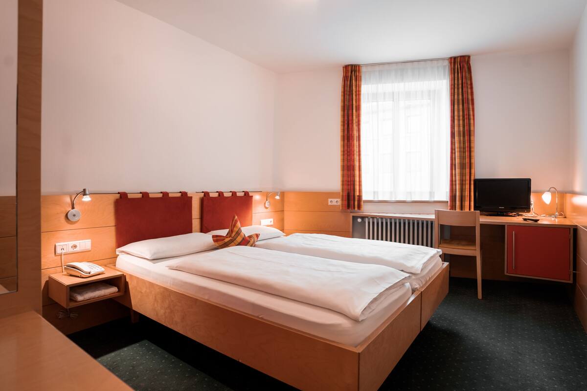 A hotel room with a bed, bedside table, lamp, TV, desk, and window with curtains.