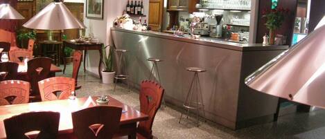 Zona de bar con taburetes, una barra y varias botellas. Se ven mesas y sillas en primer plano.