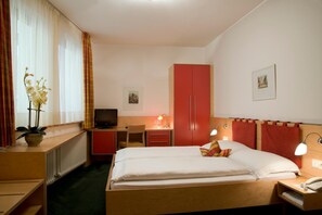 Una habitación de hotel con una cama, un escritorio con computadora, un televisor, un armario y una mesita de noche con lámpara.
