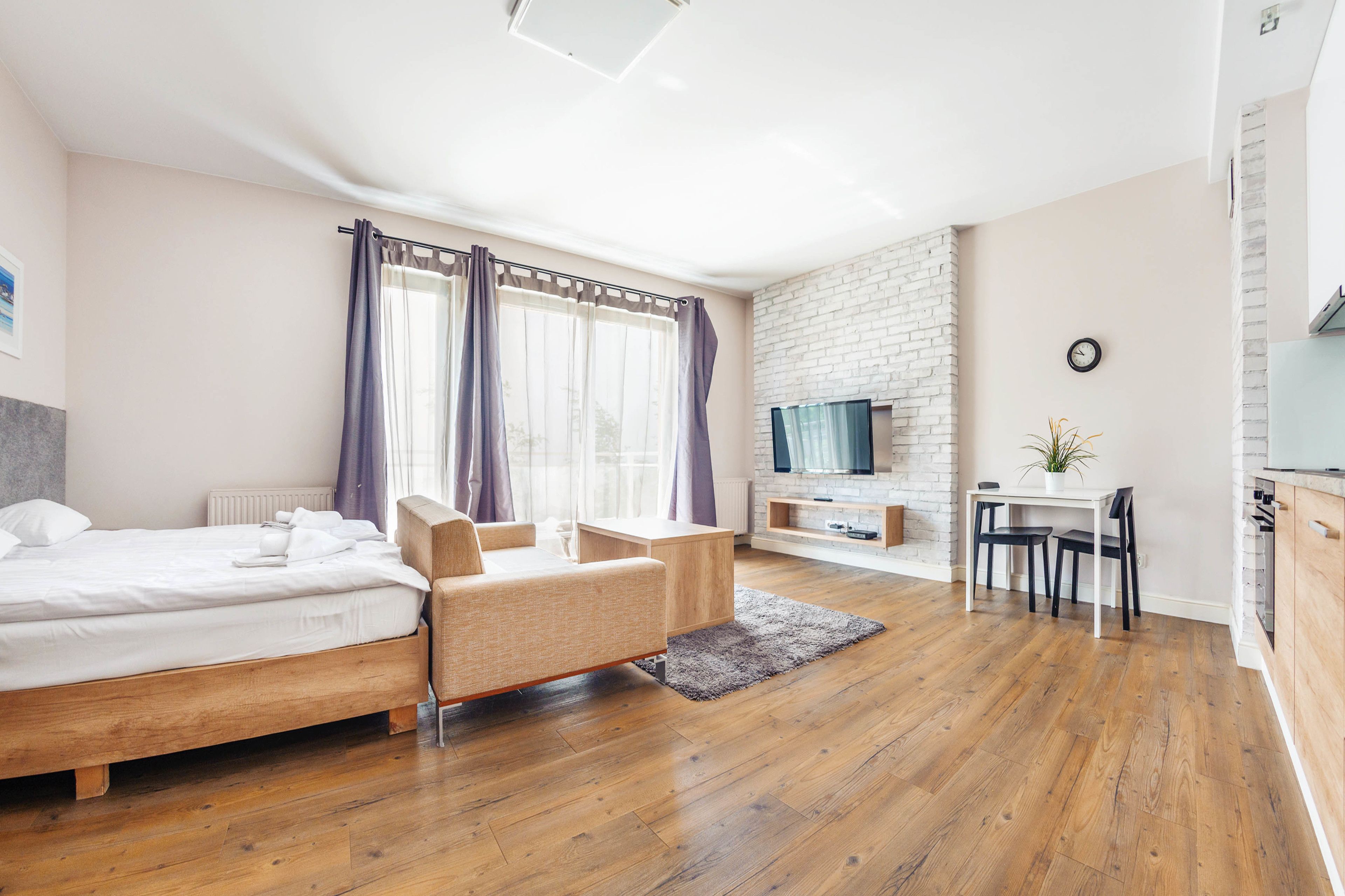 A modern hotel room with a wooden bed, a sofa, a desk with a chair, a TV, and a brick wall.
