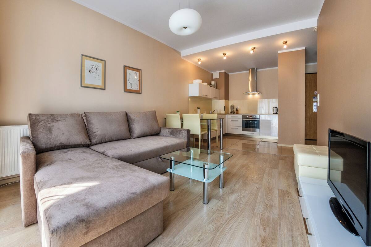 Un salon moderne avec un canapé d’angle, une table basse en verre, un téléviseur à écran plat et une cuisine en arrière-plan.