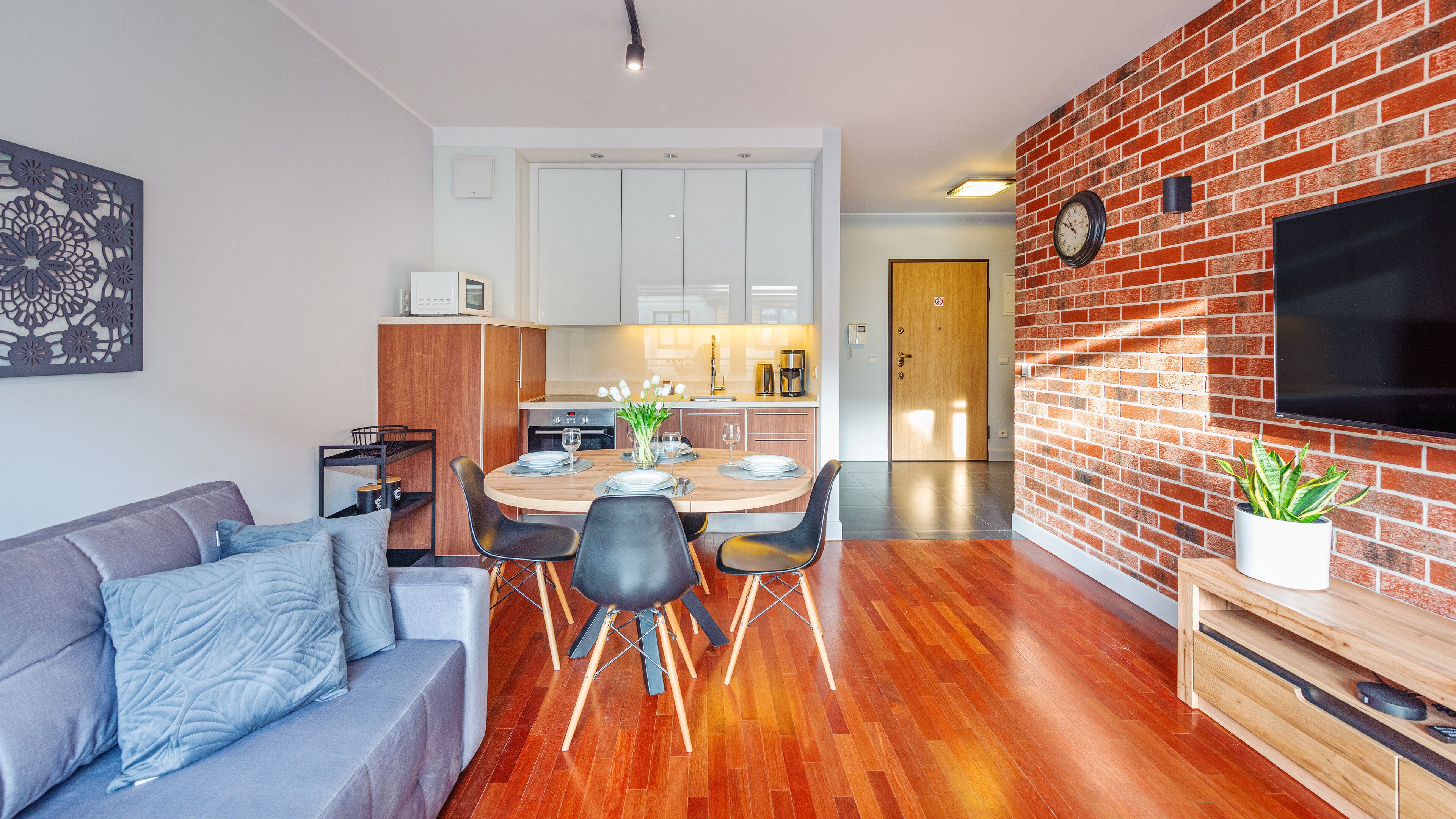 Un soggiorno moderno con un divano grigio, un tavolo da pranzo con sedie, una zona cucina con mobili bianchi e un muro in mattoni con un televisore.