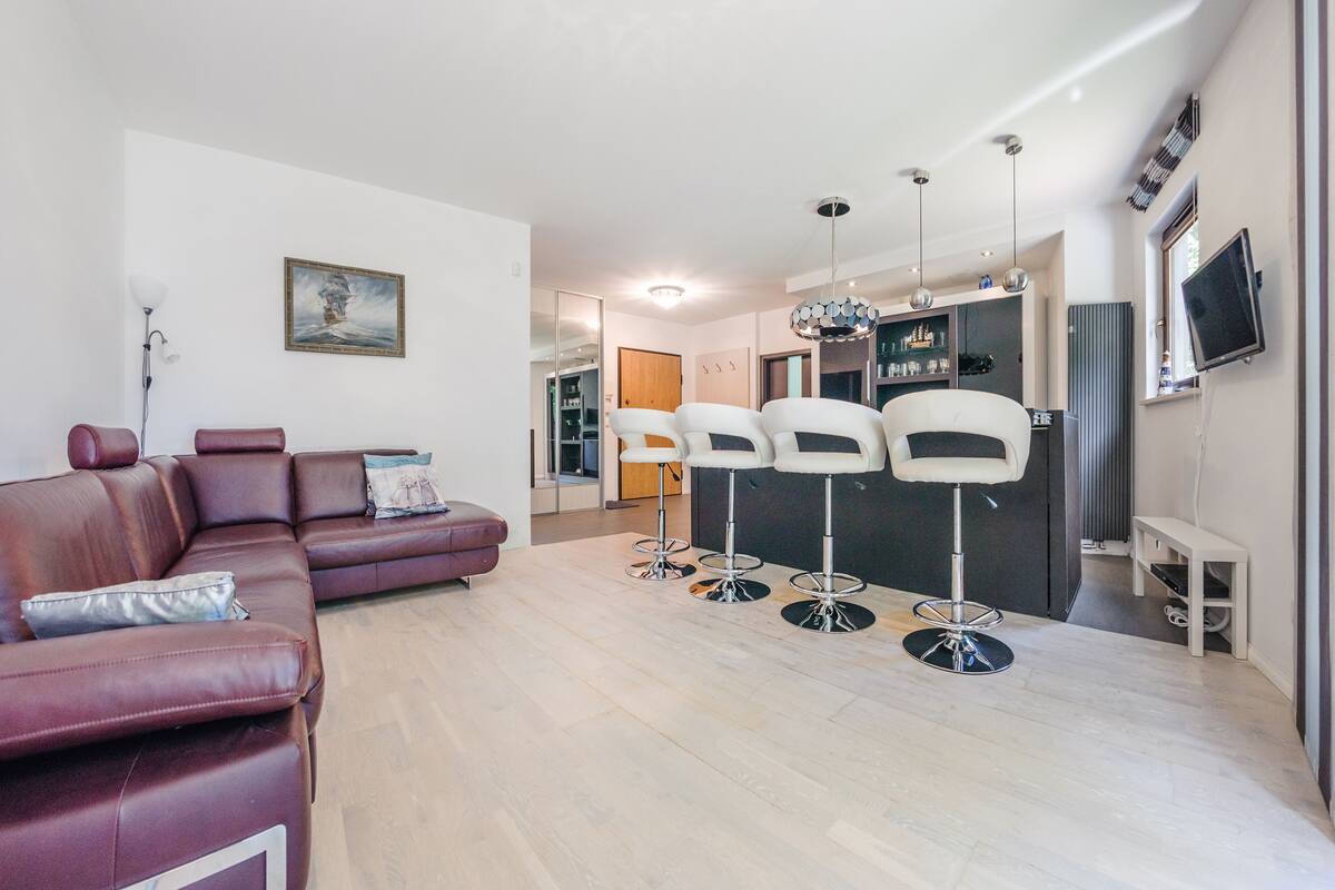 Un salon moderne avec un canapé en cuir, un coin repas doté de tabourets de bar, une cuisine avec un téléviseur et un grand miroir.