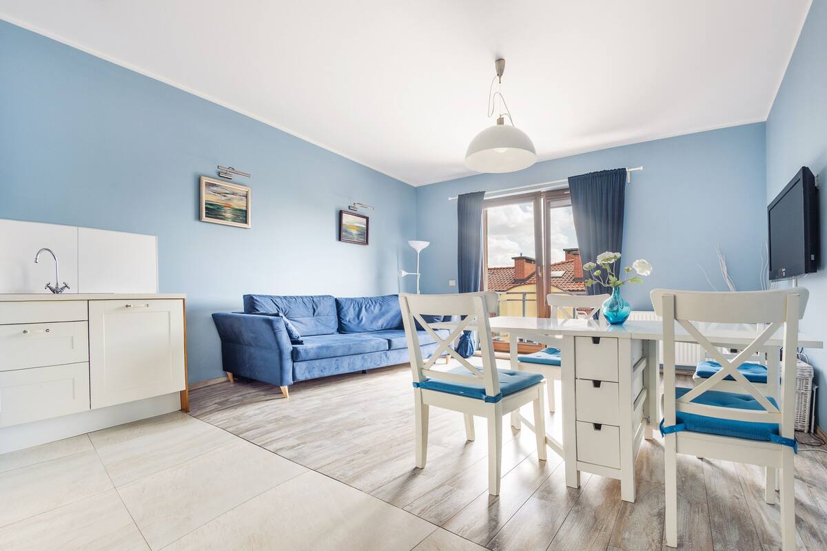 Un salon moderne avec un canapé bleu, une table à manger avec des chaises, un meuble blanc et un téléviseur fixé au mur.
