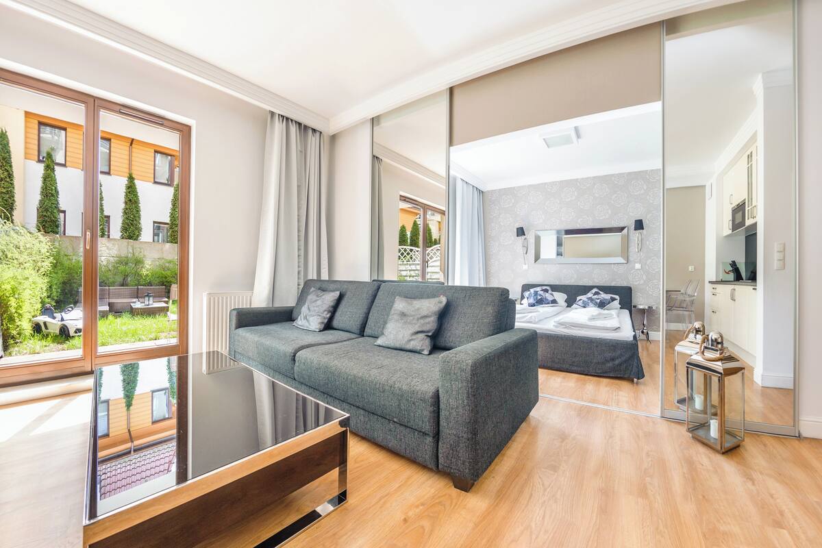 Un salon moderne avec un canapé gris, une table basse en verre et une vue sur un balcon où l’on aperçoit une voiture et de la verdure.