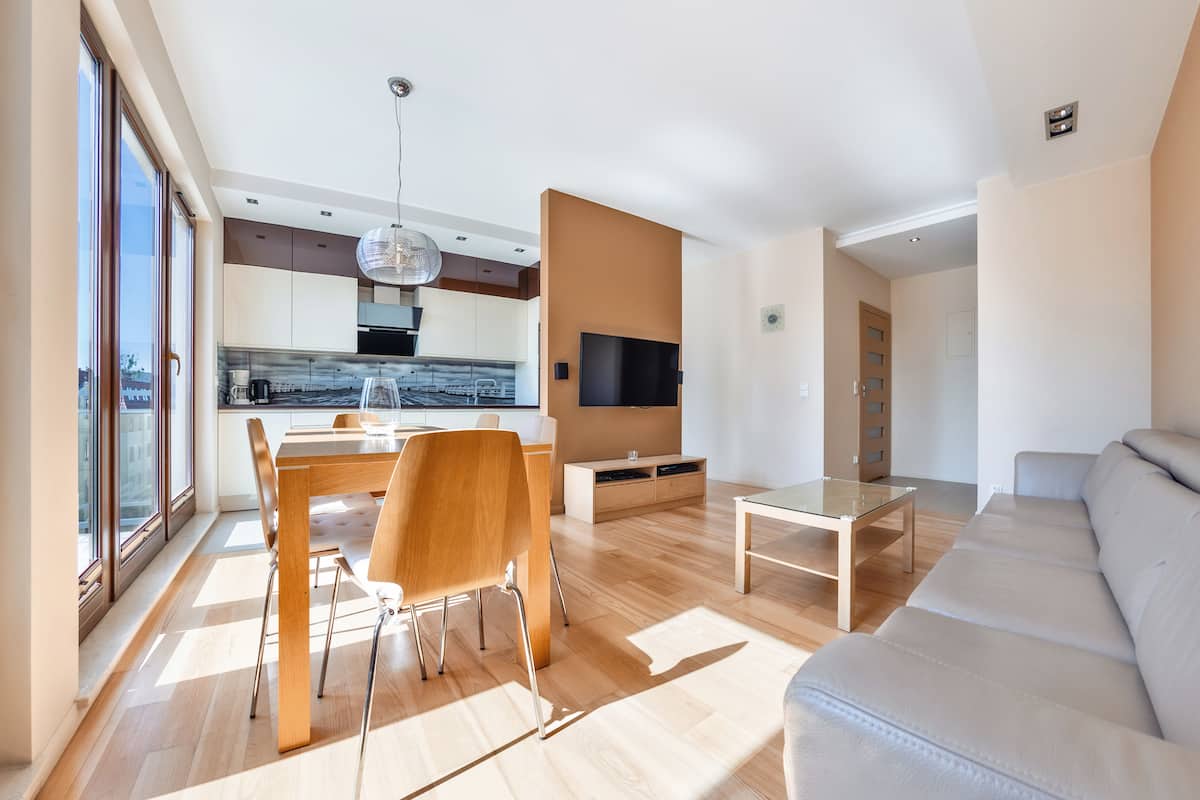 Un salon moderne comprenant un canapé, une table à manger avec des chaises, un téléviseur fixé au mur et un coin cuisine visible à travers une porte ouverte.