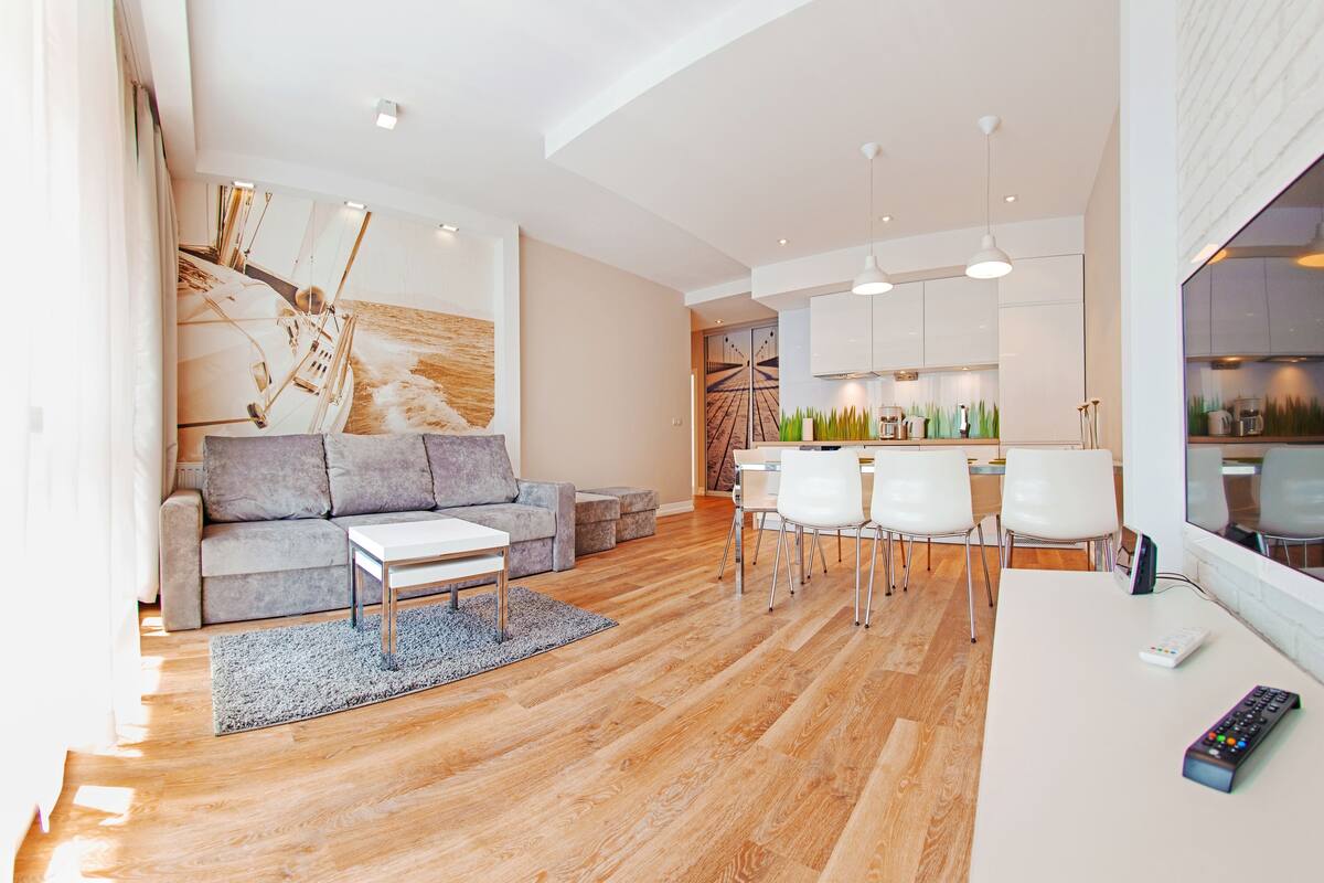 Un salon moderne avec un canapé, une table basse et un coin repas avec des chaises. On aperçoit au fond une cuisine équipée de placards et d’une table à manger.