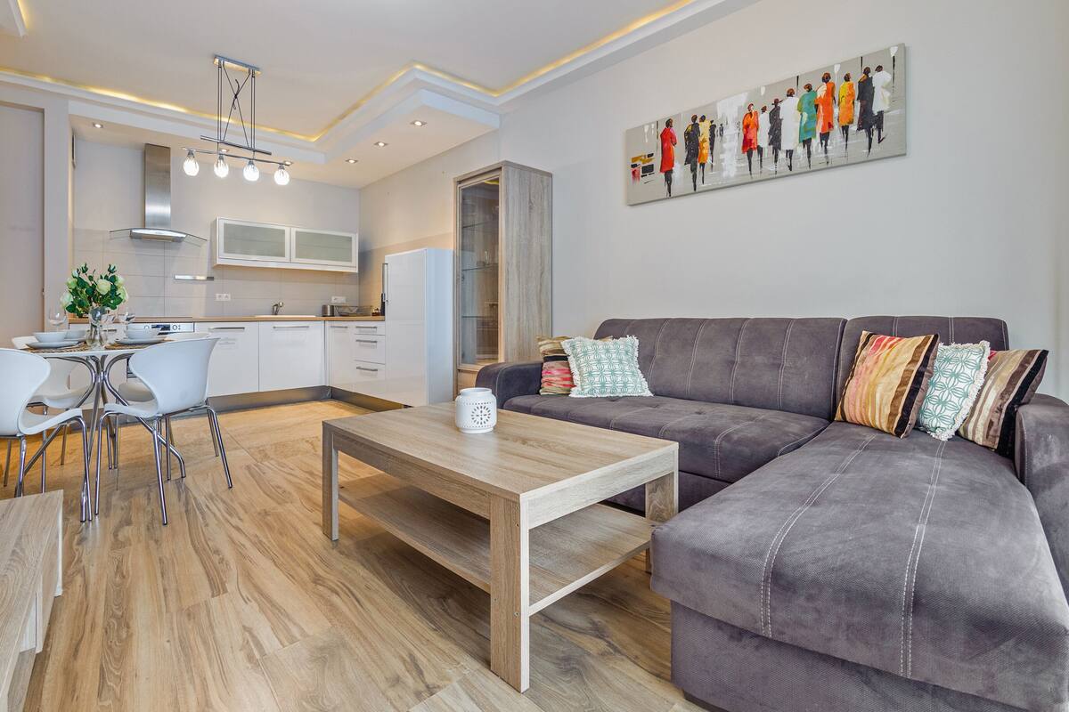 Un salon moderne avec un canapé gris, une table basse en bois et des chaises blanches. On aperçoit une cuisine avec des meubles blancs et un coin repas comprenant une table et des chaises.