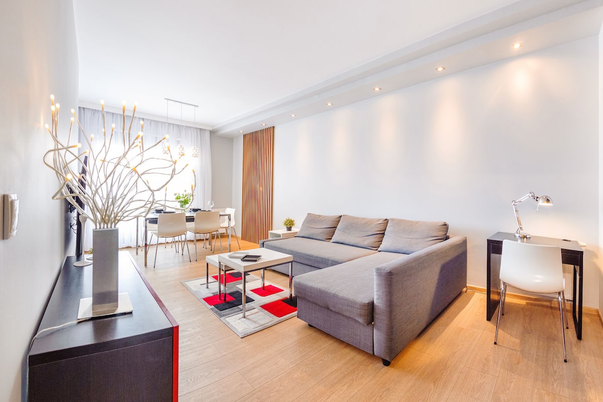 Un salon moderne avec un canapé gris, une table à manger et une table basse. On y trouve une petite table d’appoint avec une lampe, ainsi qu’un vase décoratif orné de branches.