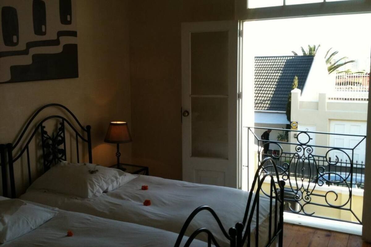 Habitación de hotel con cama, mesita de noche, lámpara y balcón con barandilla.