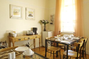A dining area with a wooden table set for four, a small refrigerator, and a coffee maker on a wooden table. - Cheviot Place Guest House (Cape Town)