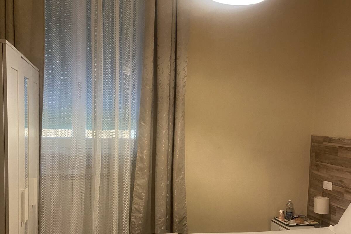 A hotel room with a bed, a nightstand, a lamp, and a window with sheer curtains.