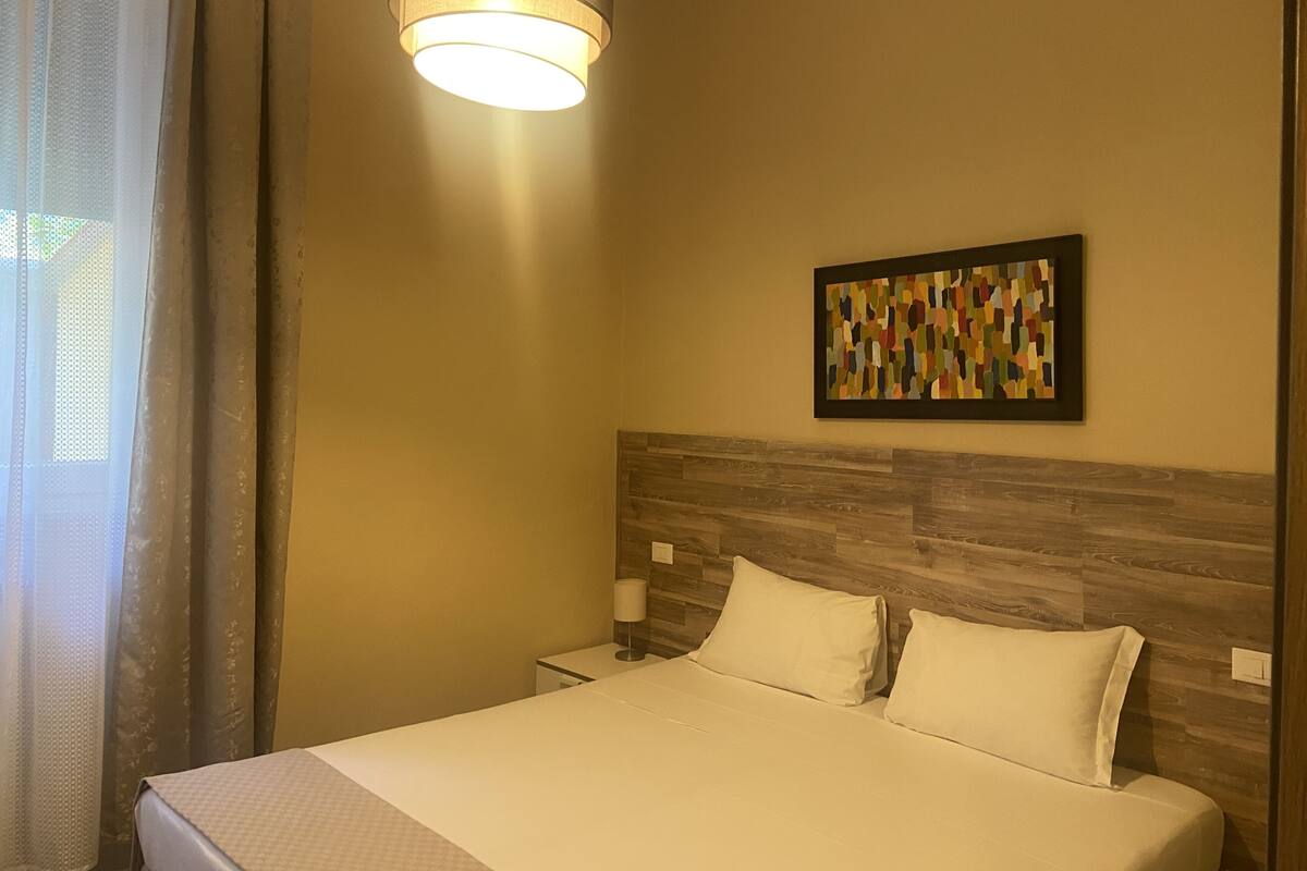 A hotel room with a bed, a nightstand, a lamp, a wall-mounted artwork, and a window with curtains.